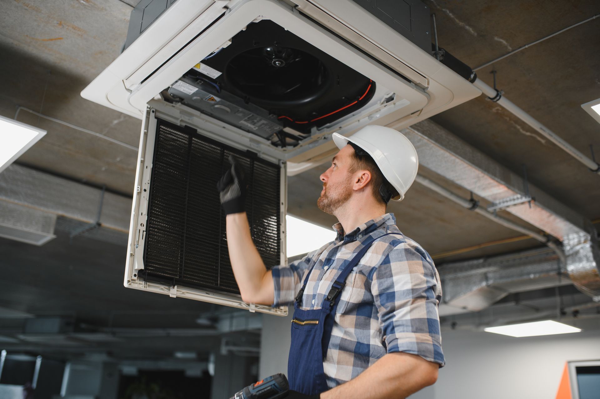 HVAC technician inspecting ceiling-mounted air conditioning unit, wearing a hard hat, in a commercial space.