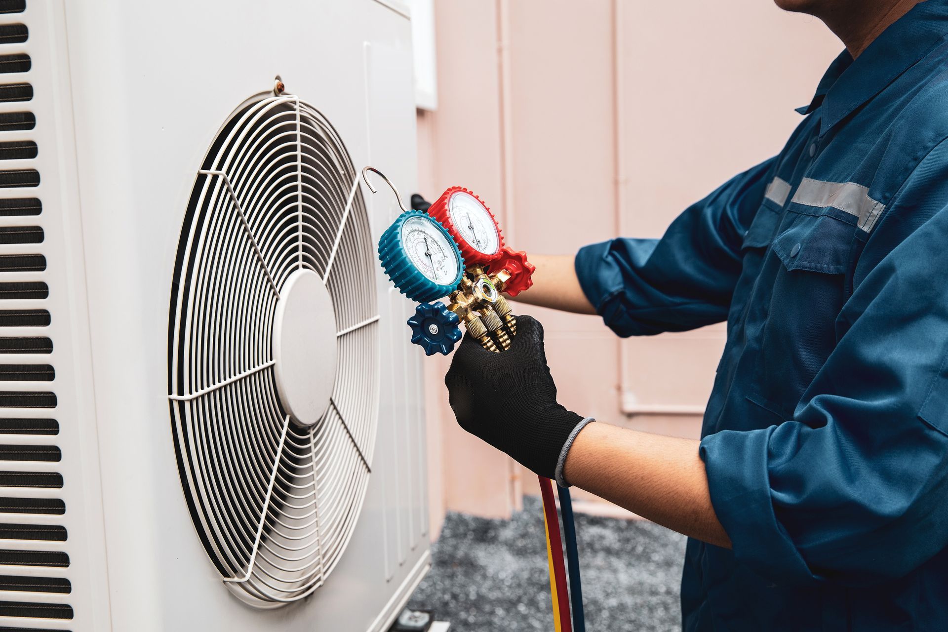 HVAC technician in blue overalls servicing an outdoor AC unit with gauges; black gloves.