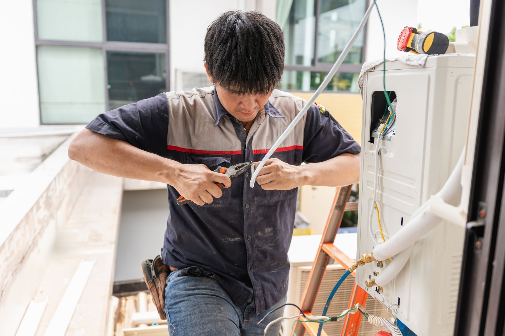 HVAC technician in a blue shirt and cap, inspecting and repairing electrical components of a unit, indoors.