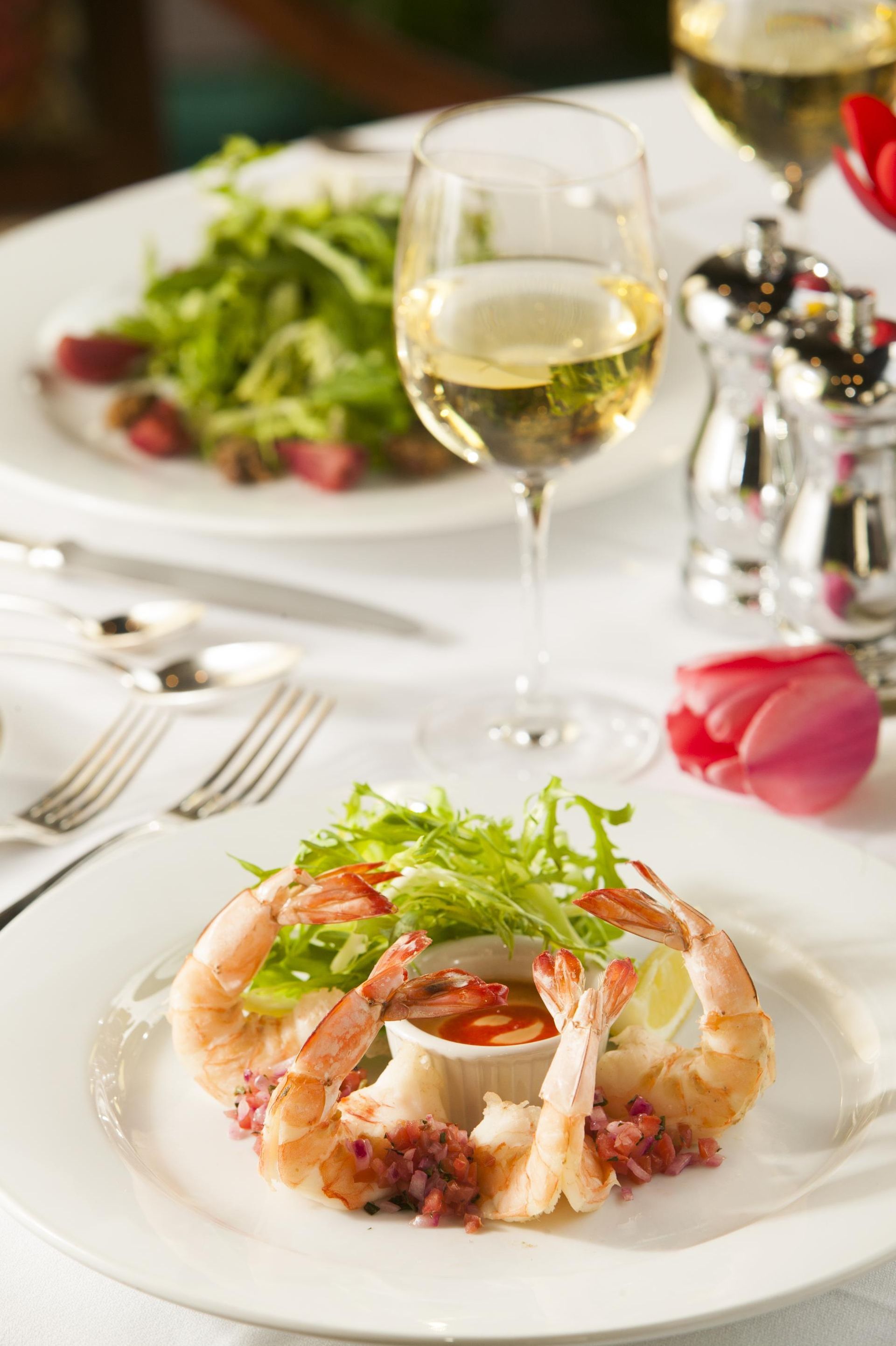 Elegant seafood dish with greens on a white table, beside a glass of white wine and pink rose petals