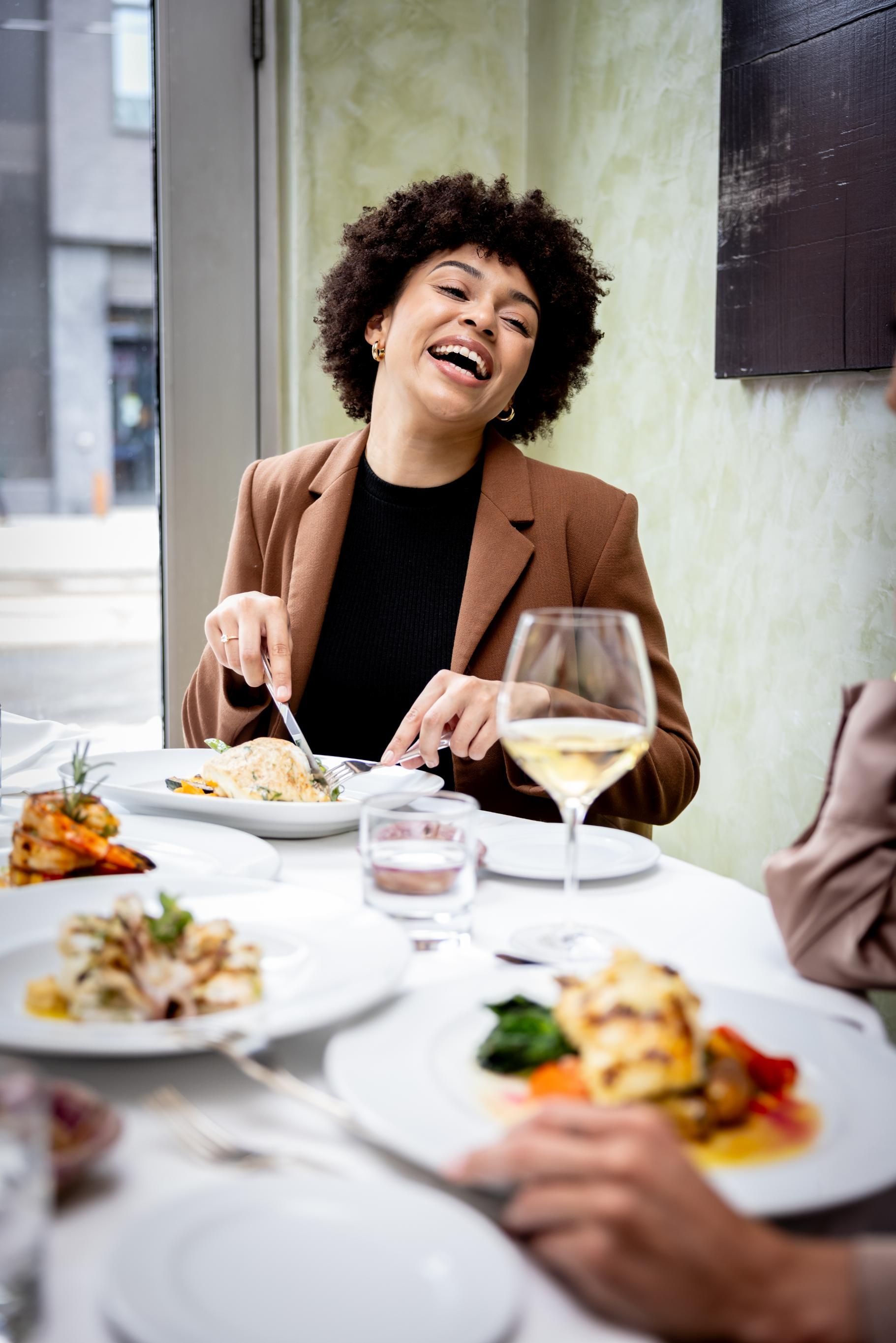 Person laughing at a restaurant table with plated dishes and wine glasses