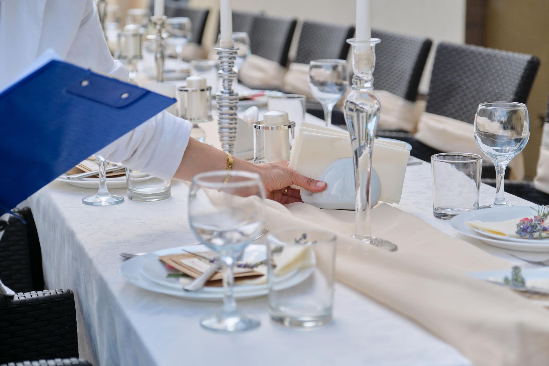 Elegant dining table being set with white tablecloth, glassware, and plates; a person adjusts a place setting.