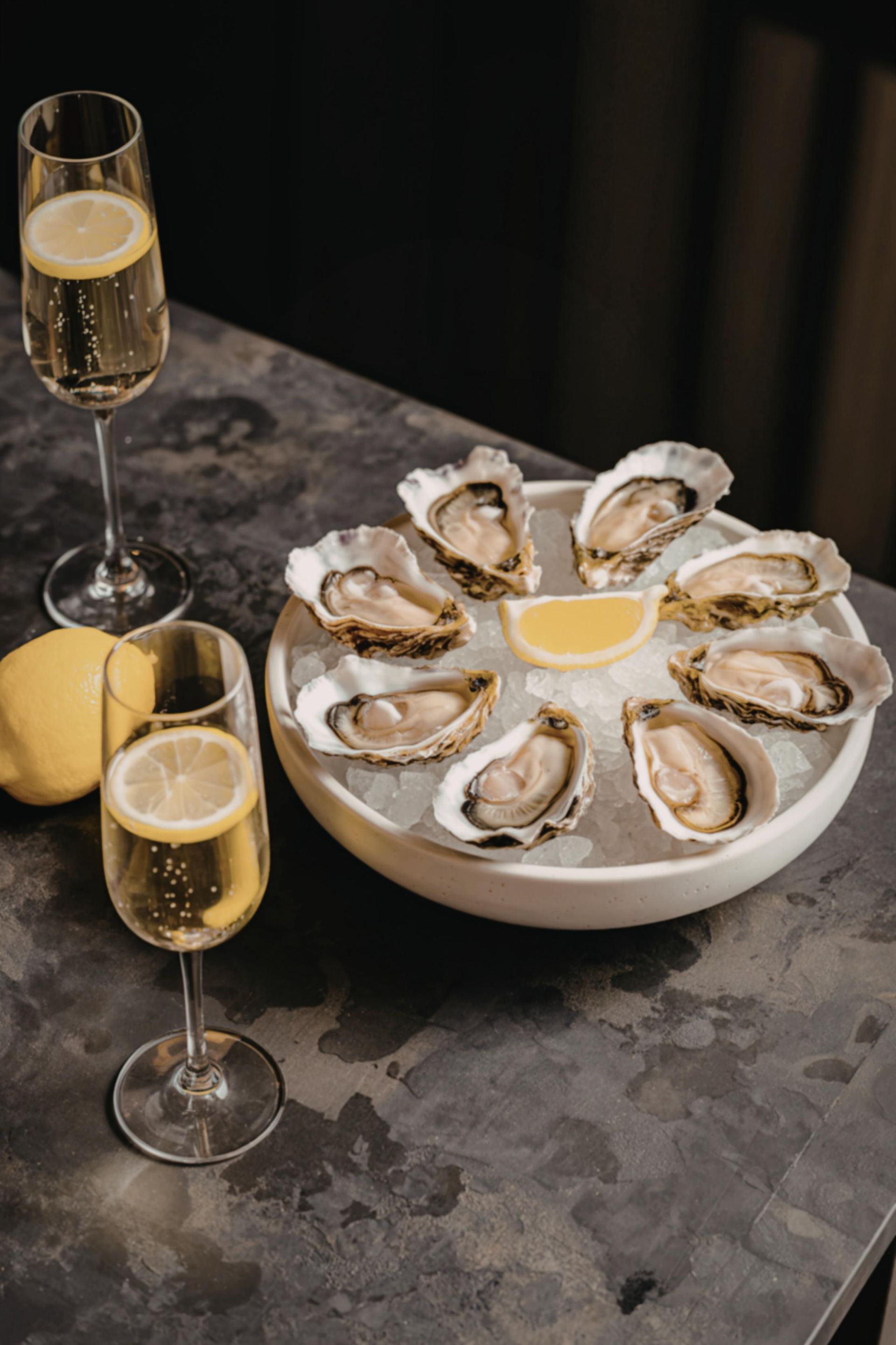 Oysters on ice with lemon wedges and two glasses of champagne on a dark stone table