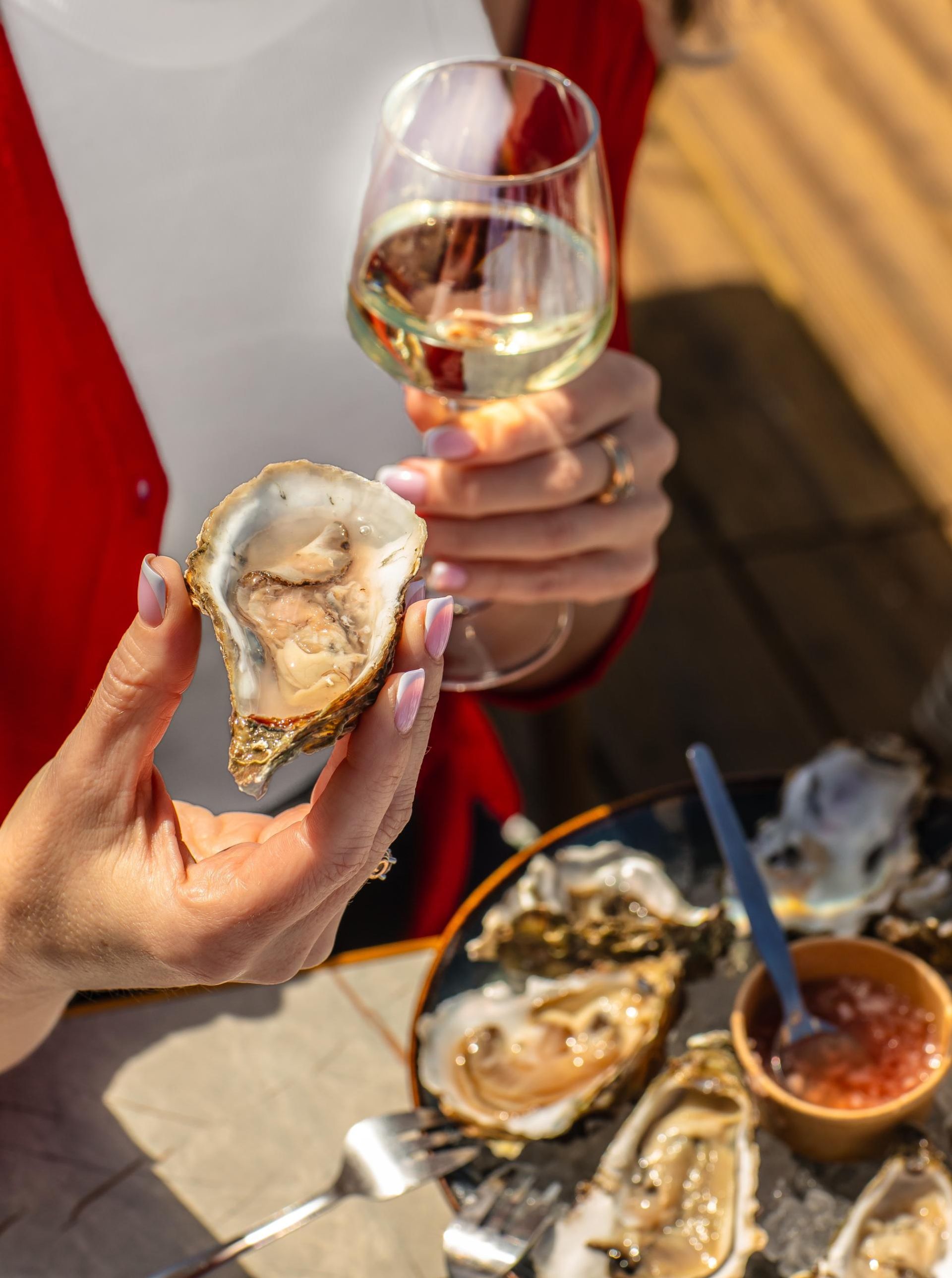 Hands holding oysters and a glass of white wine at a sunlit seafood table