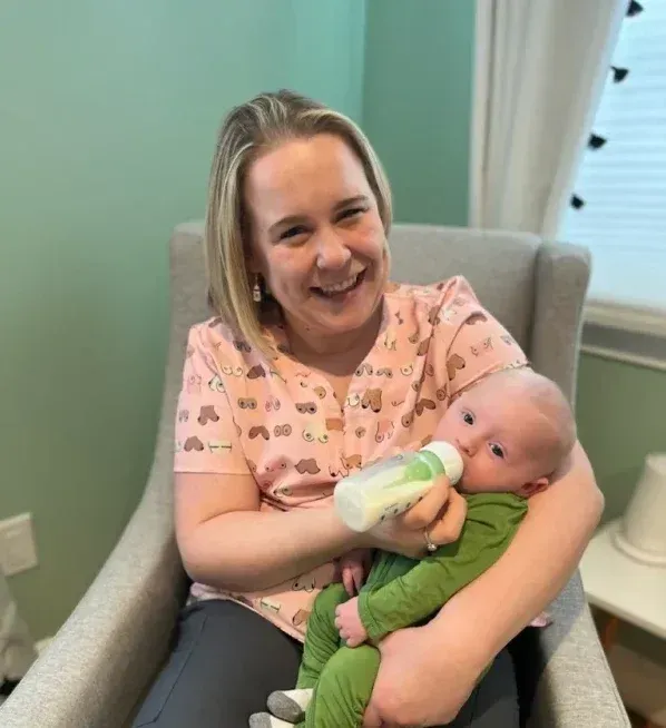 Woman in pink shirt feeds a baby in a green onesie a bottle while smiling in a gray chair.
