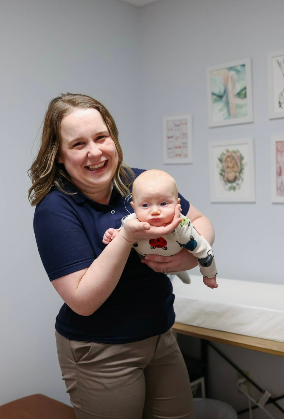 Woman holding a baby in an office, smiling. Baby in a onesie, looking at the camera. Neutral background, artwork.