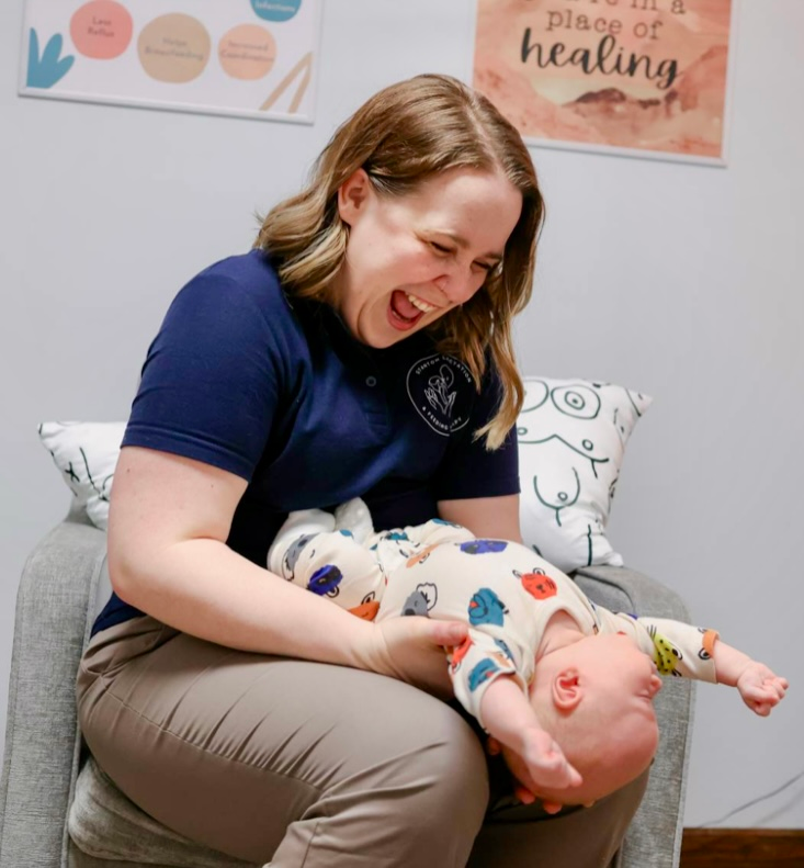 Woman laughing, holding infant in her lap. Setting appears to be an office.