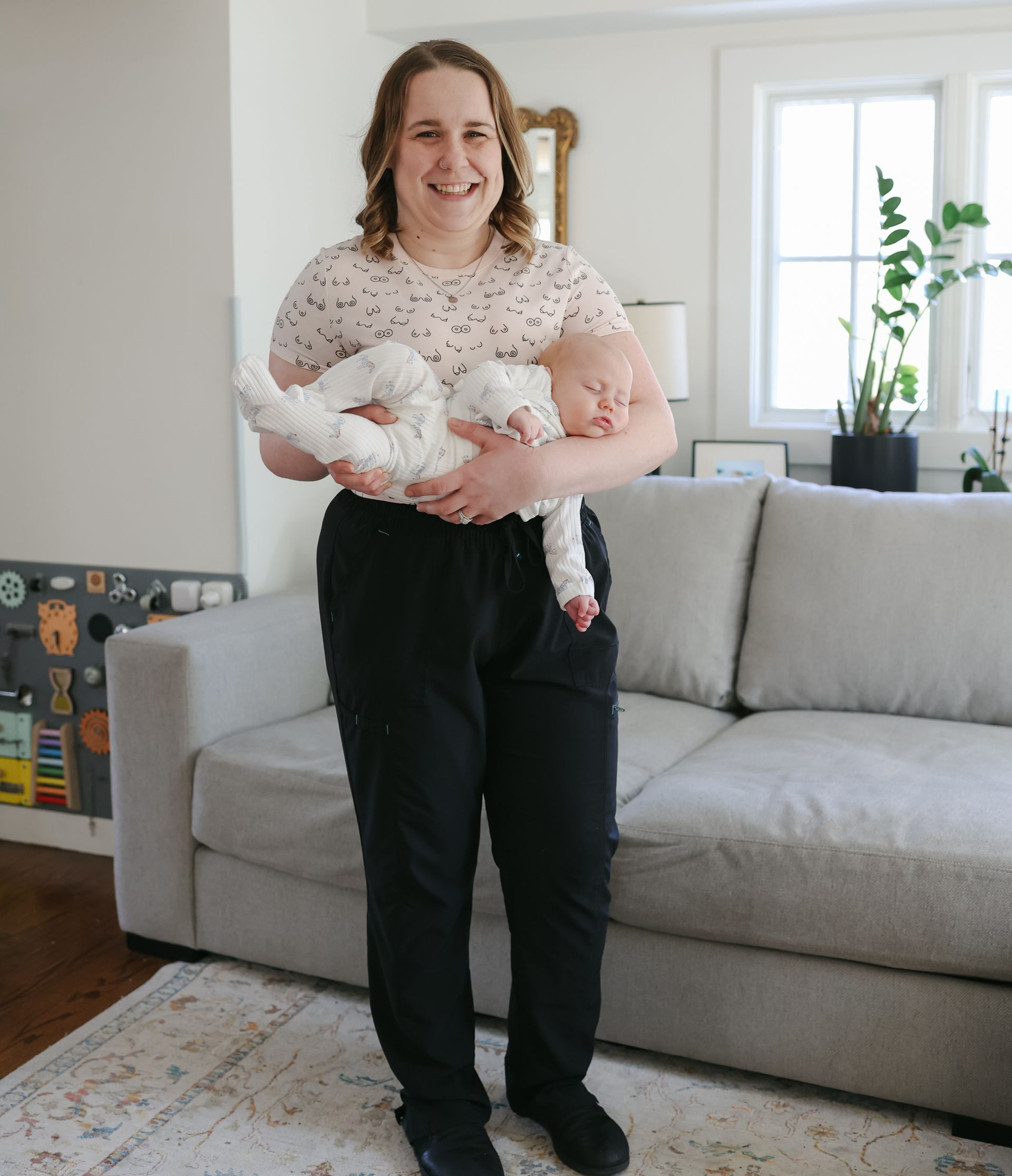 Woman holding a baby indoors, smiling. She wears a patterned shirt, black pants, and holds the sleeping baby.