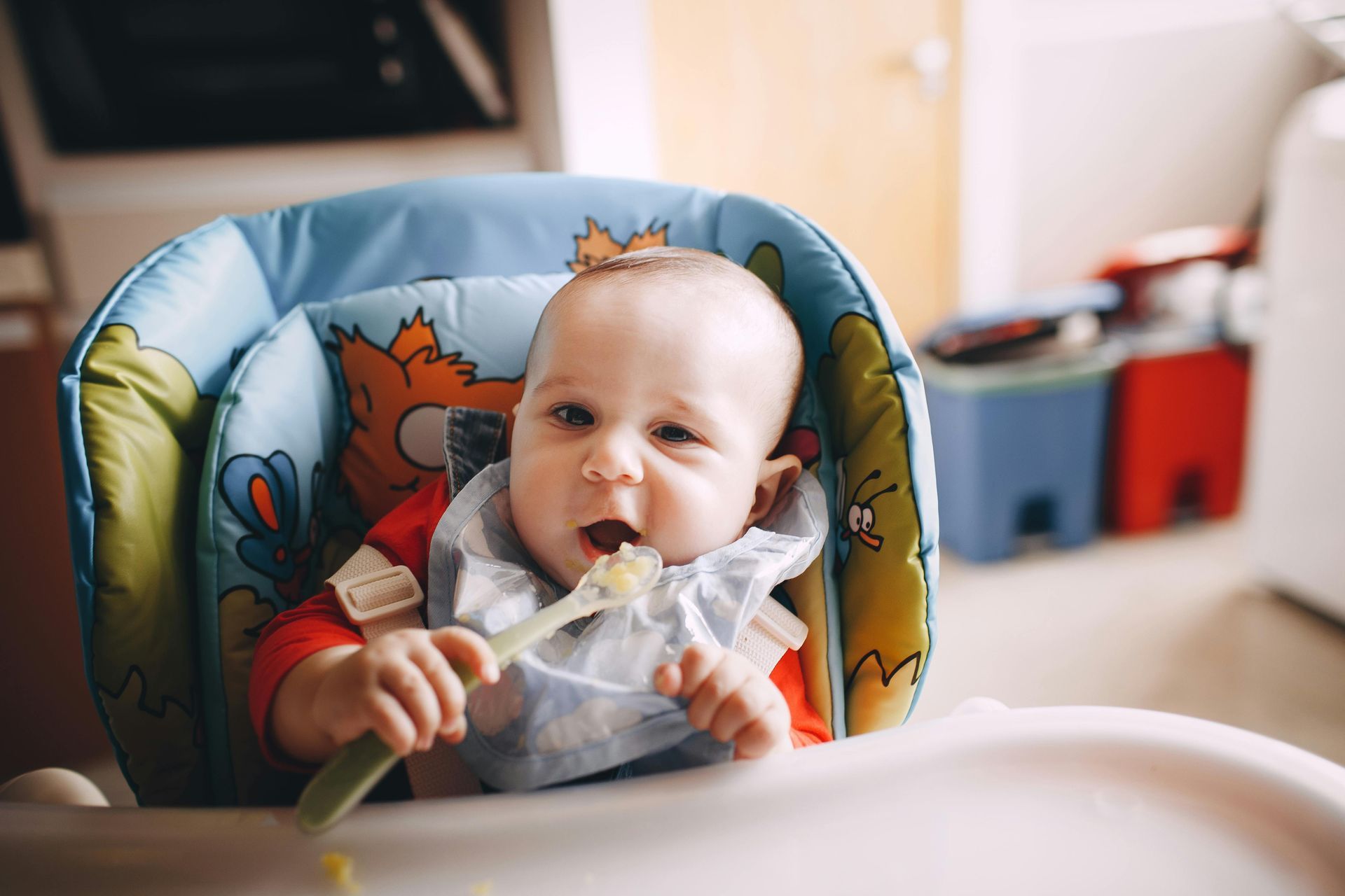 Baby in a blue high chair, wearing a bib, eating with a spoon, looking at the camera.