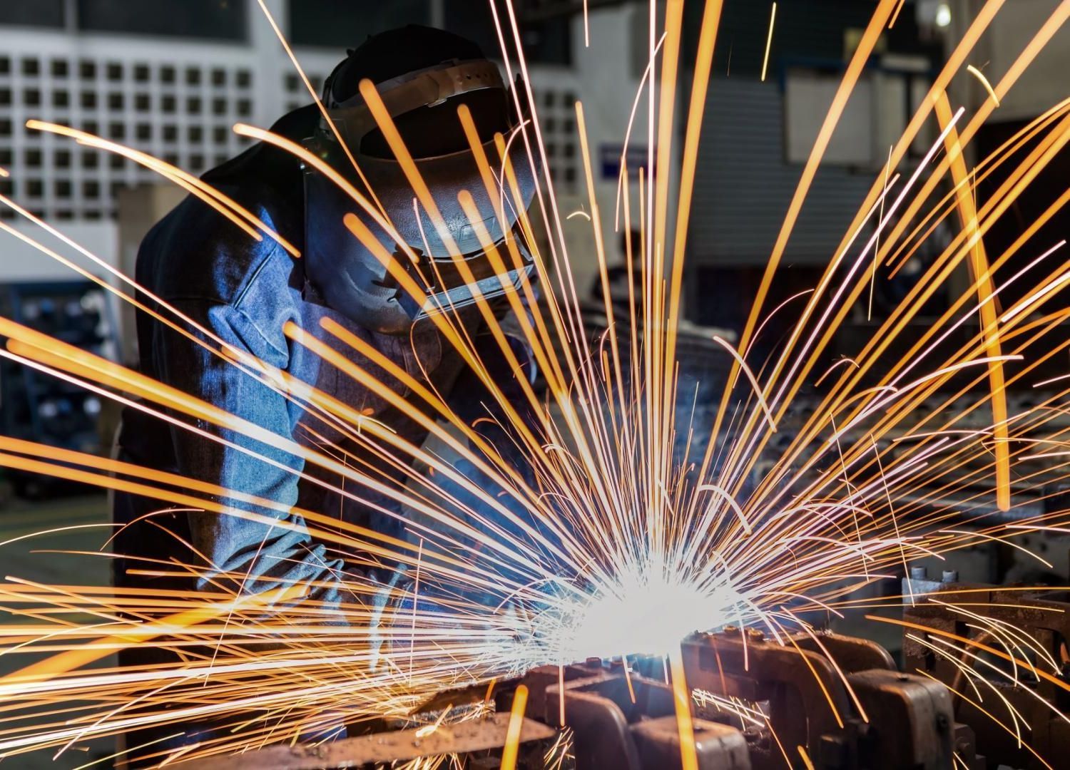 Soudeur en veste bleue produisant des étincelles brillantes lors de travaux de métallurgie dans un atelier