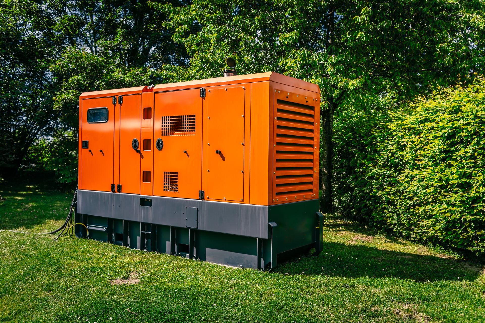Orange and gray industrial generator in a grassy area, with green bushes and trees in the background.