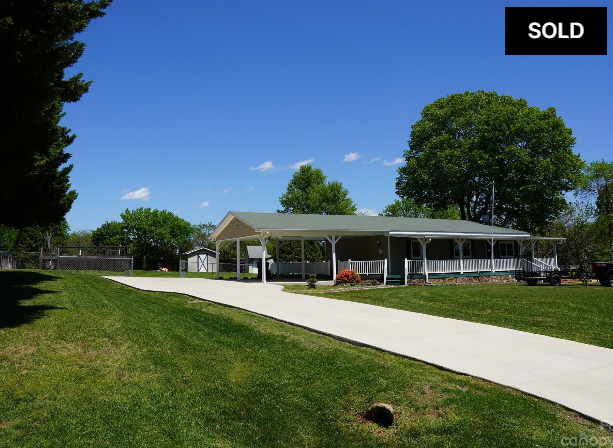 Modern single-story building with a white roof, green lawn, and driveway under a blue sky; sold sign in corner