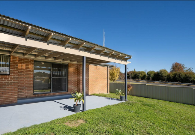 Brick house with a covered patio, pots of plants, and a green lawn under a blue sky.