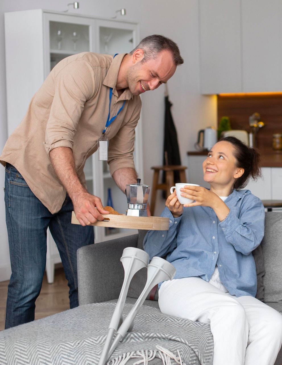 Man serves woman breakfast in living room; woman smiles, crutches near.