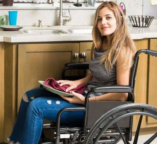 Woman assisting a young girl in a wheelchair, wiping her face with a washcloth. Bathroom setting.