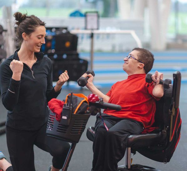 Woman coaches a man in a wheelchair using dumbbells in a gym. Both have excited expressions.