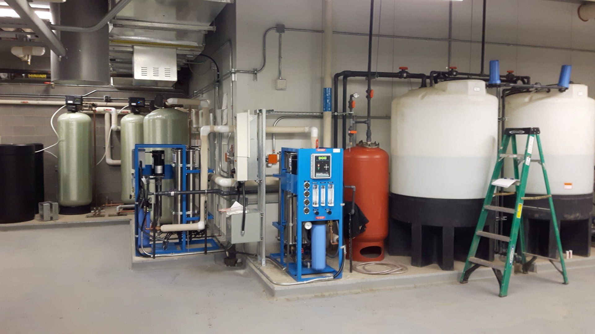 Industrial utility room with blue equipment tanks, large white and red vessels, pipes, and a green ladder.