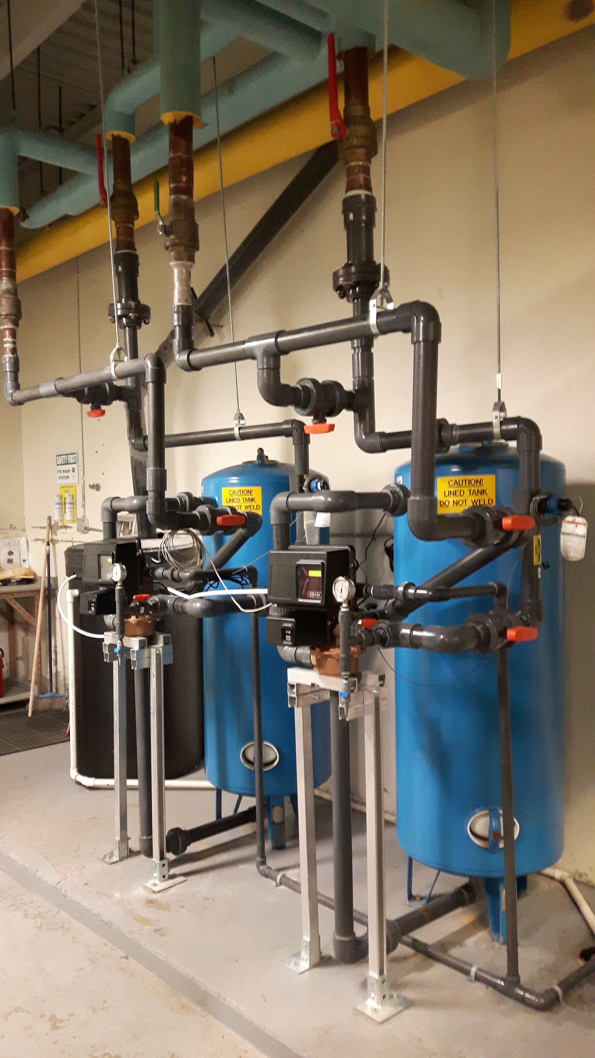 Industrial filtration tanks and piping in a utility room, with blue and black equipment.
