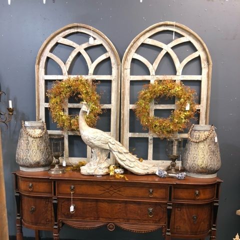 White peacock figurine on a wooden sideboard, flanked by wreaths and vintage arched windows against a gray wall