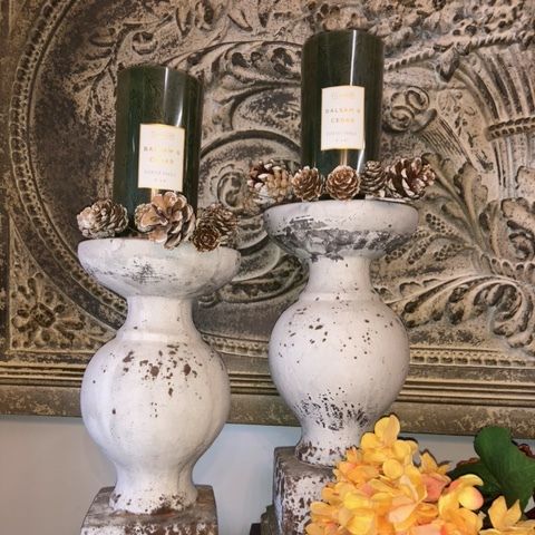 Two distressed white candle holders with pinecones and green candles on a decorative wall hanging.