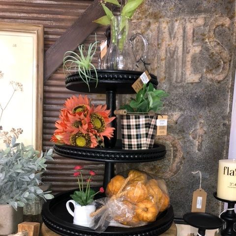 Tiered black display shelf with flowers, potted plants, pumpkins, and kitchen decor in a rustic room