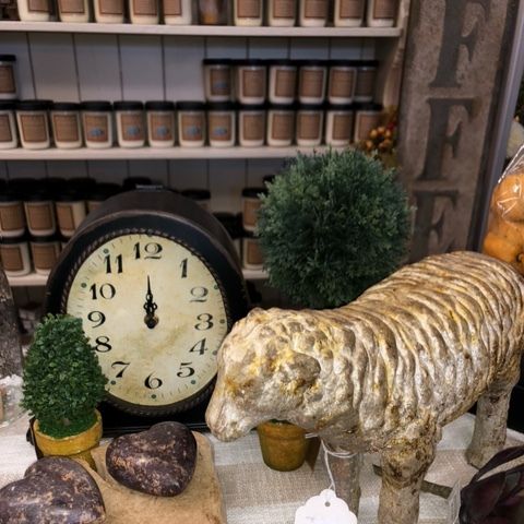 Vintage display with an old clock, small potted trees, heart-shaped stones, and a sheep figurine in front of shelves