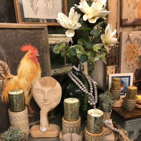 Decorative tabletop display with white flowers, candles, a carved face, a rooster figurine, and framed art