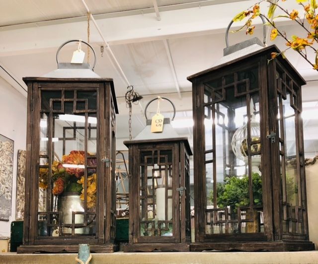 Three dark wooden lanterns with glass panes and small plants inside, displayed on a shelf outdoors.