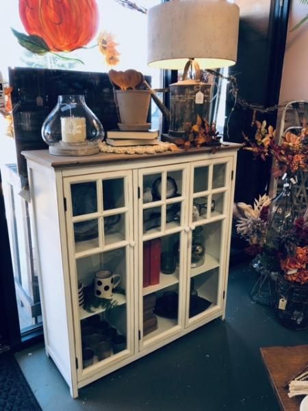 White glass-front cabinet with decor and a lamp in a cozy room, beside a floral arrangement.