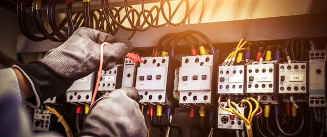 Gloved hands adjusting electrical circuit breakers in a panel with tangled wires and a warm backlight