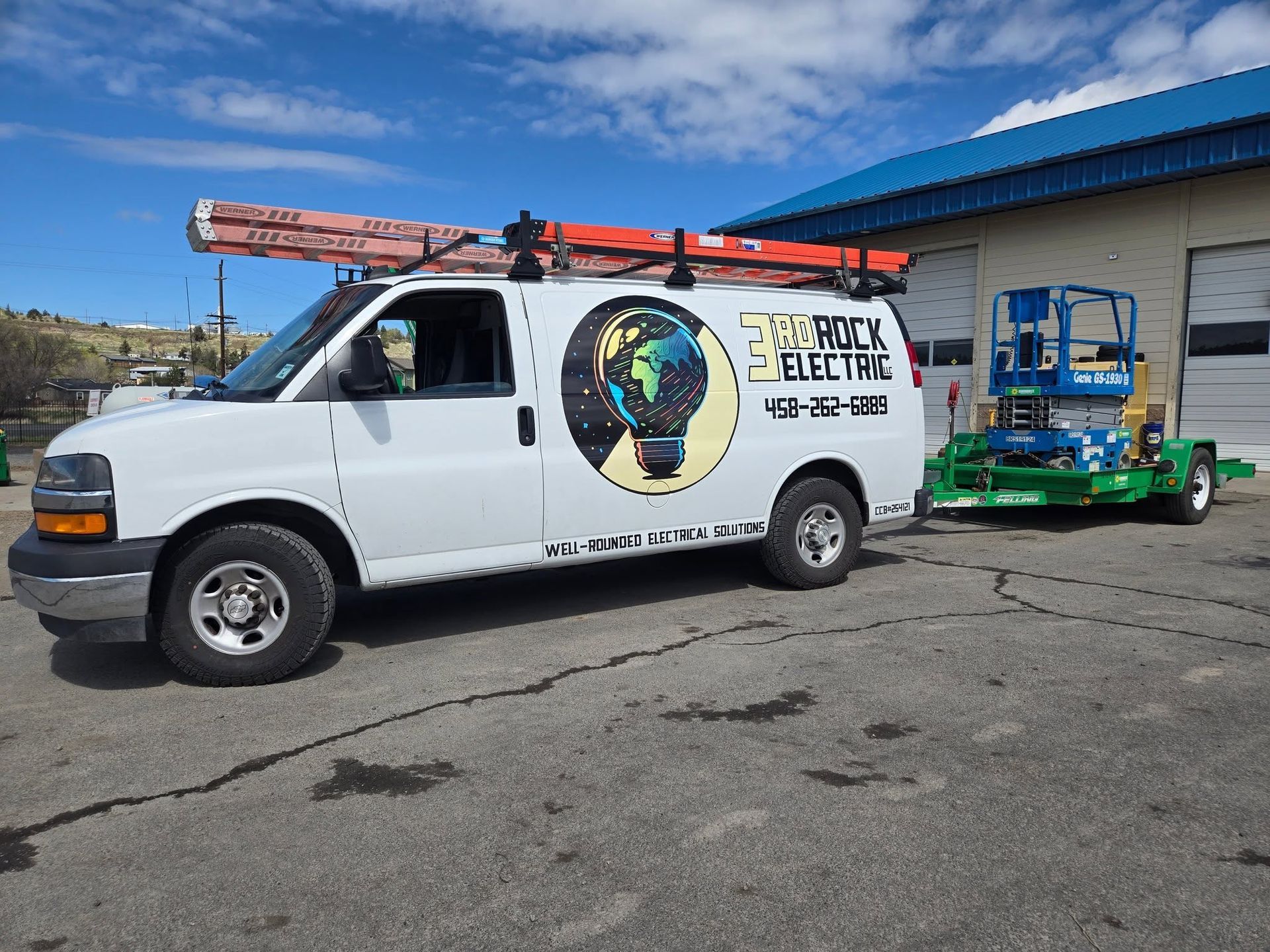 White utility van with ladder rack parked beside a blue-roofed building, towing green equipment.