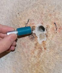 Hand holding a blue flashlight near a small hole in a concrete wall with peeling paint
