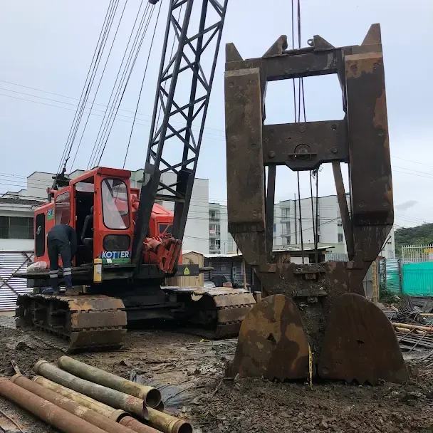 Excavadora roja sobre orugas con garra en una obra en construcción embarrada junto a tuberías y una estructura de acero.