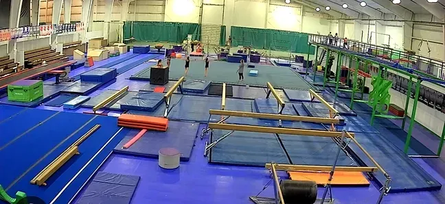 Indoor gymnastics gym with blue mats, trampolines, balance beams, and a few people in the background