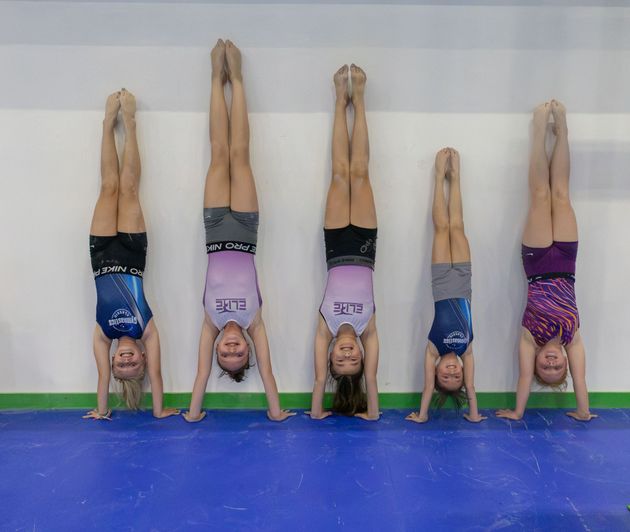 Seven gymnasts doing handstands against a wall on a blue mat, wearing colorful leotards.