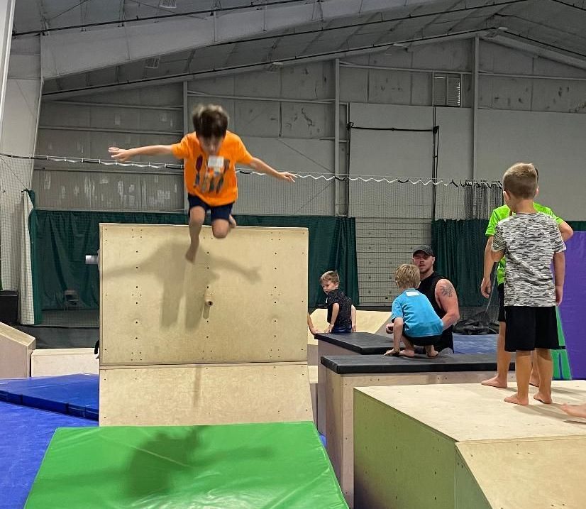 Child in orange shirt jumping off a foam block in an indoor gym, with other children nearby.