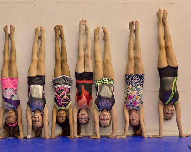 Seven gymnasts doing handstands against a wall on a blue mat, wearing colorful leotards.
