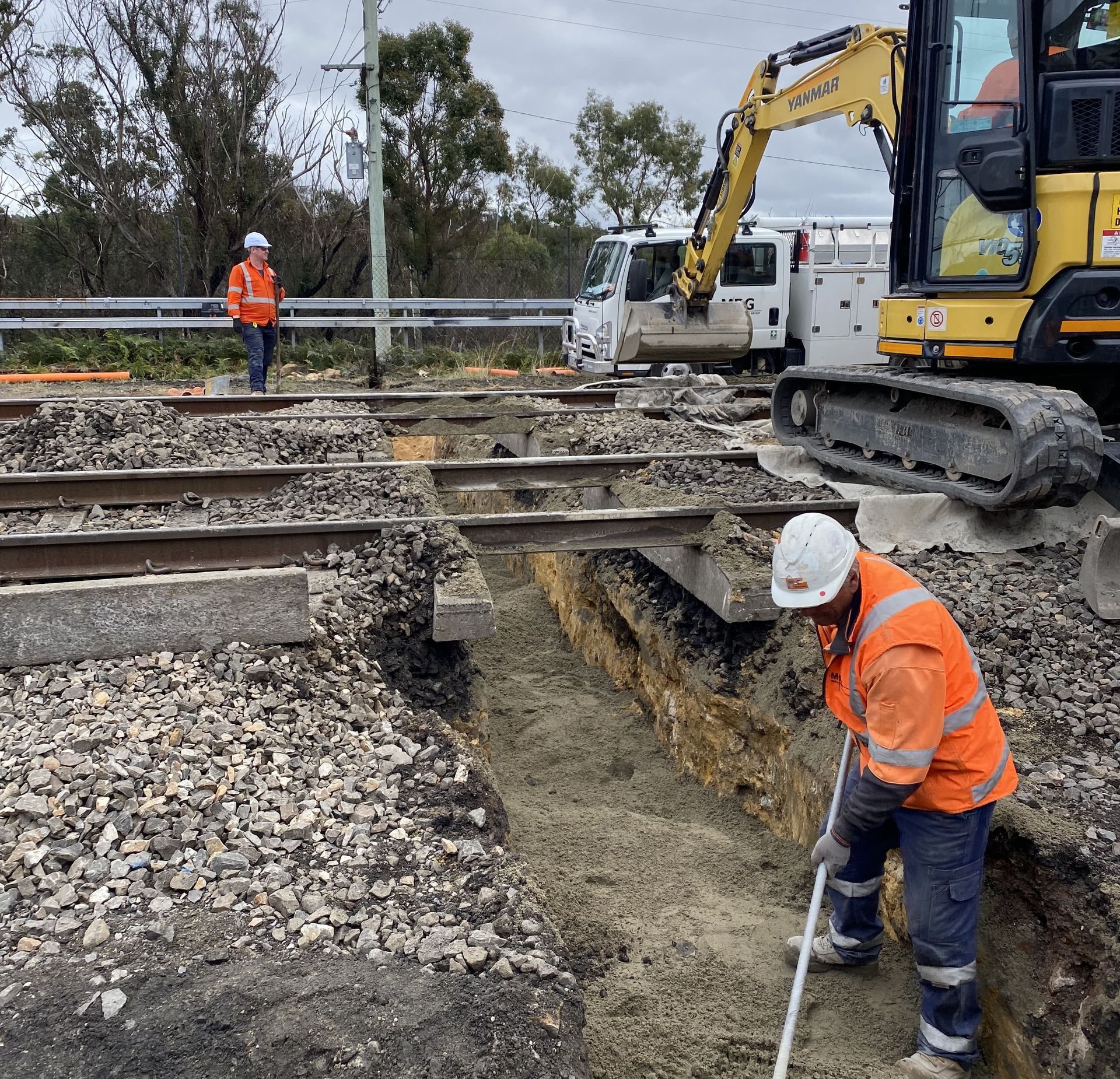 A man in an orange vest is standing in a trench next to a yellow excavator