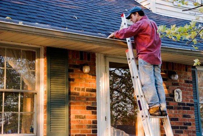 Worker on a ladder painting a brick house exterior near the roofline