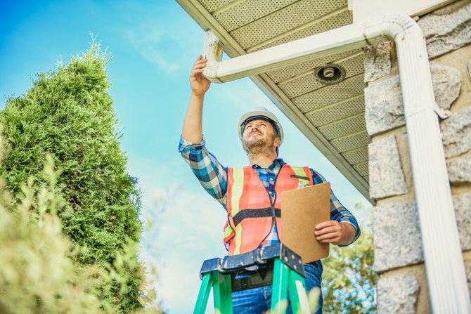 Contractor inspecting a house roof with clipboard, reaching up under the eaves outside.