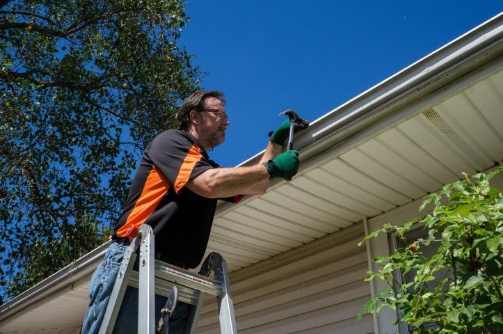 Man using a ladder and drill to install siding under a roof eave on a house