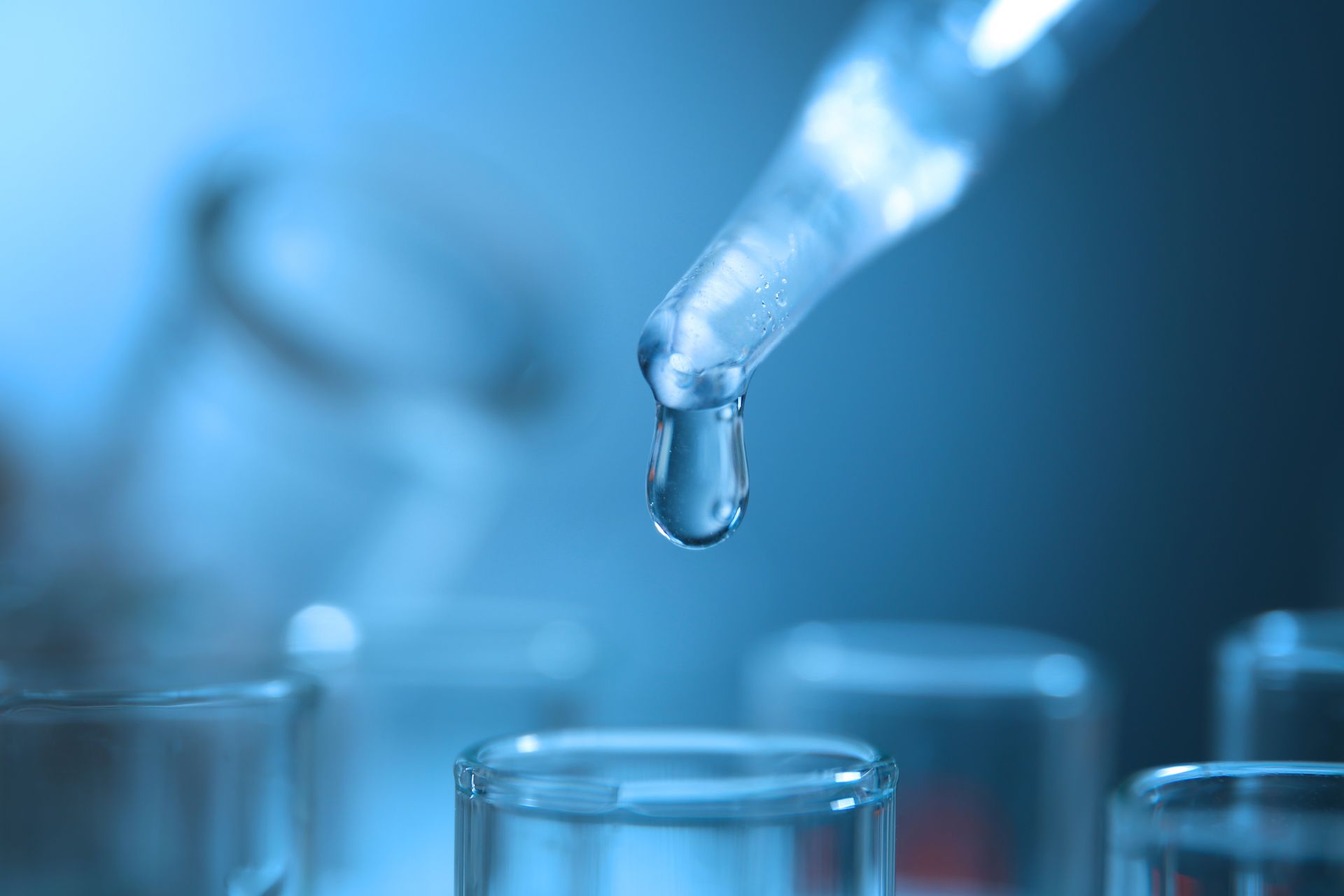 Droplet of liquid falling from a pipette into a test tube in a blue-toned lab setting.