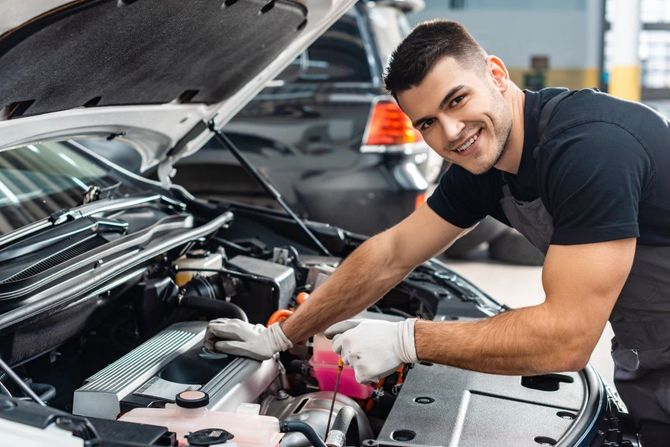 Mechanic smiling while working under an open car hood in an auto repair shop