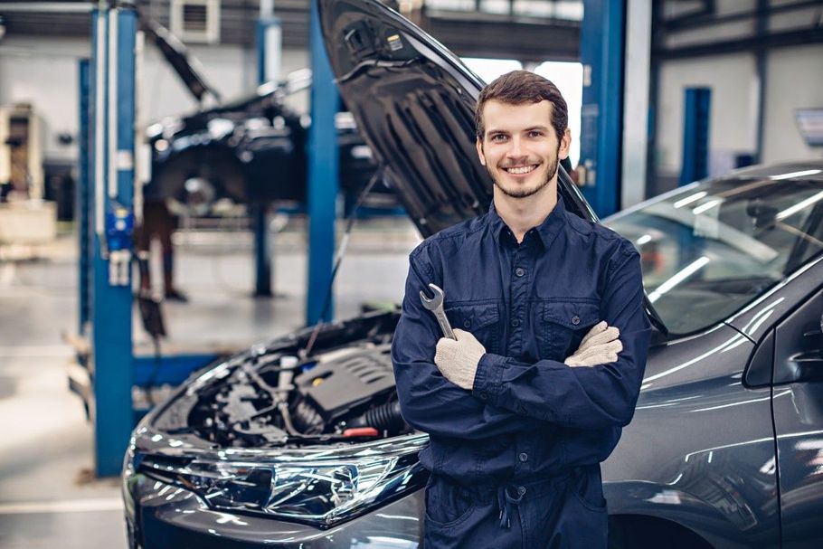 Mechanic in blue coveralls standing with arms crossed beside a car in an auto repair shop.