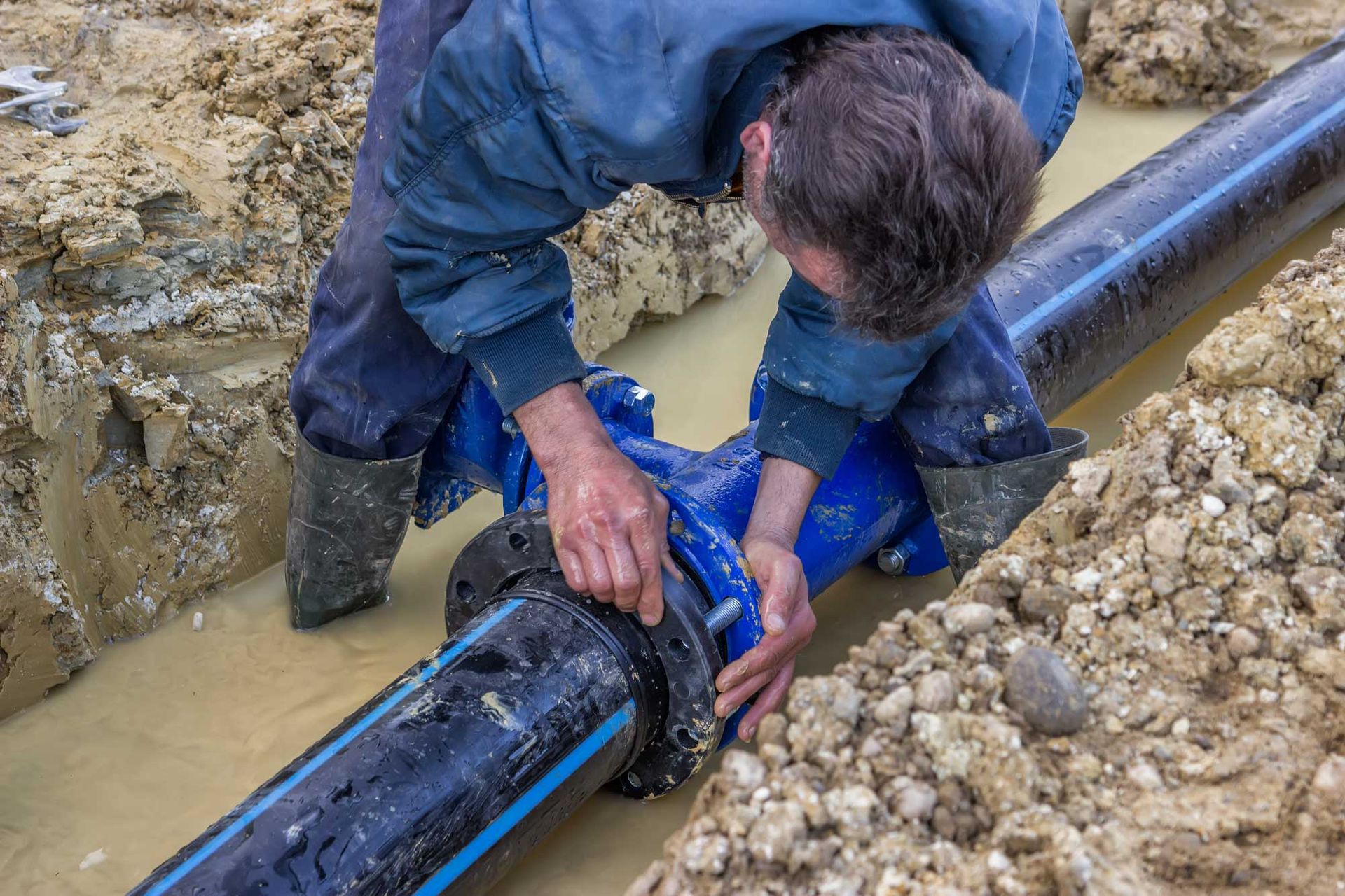 Worker installing or repairing a large water pipe connection in a muddy trench.