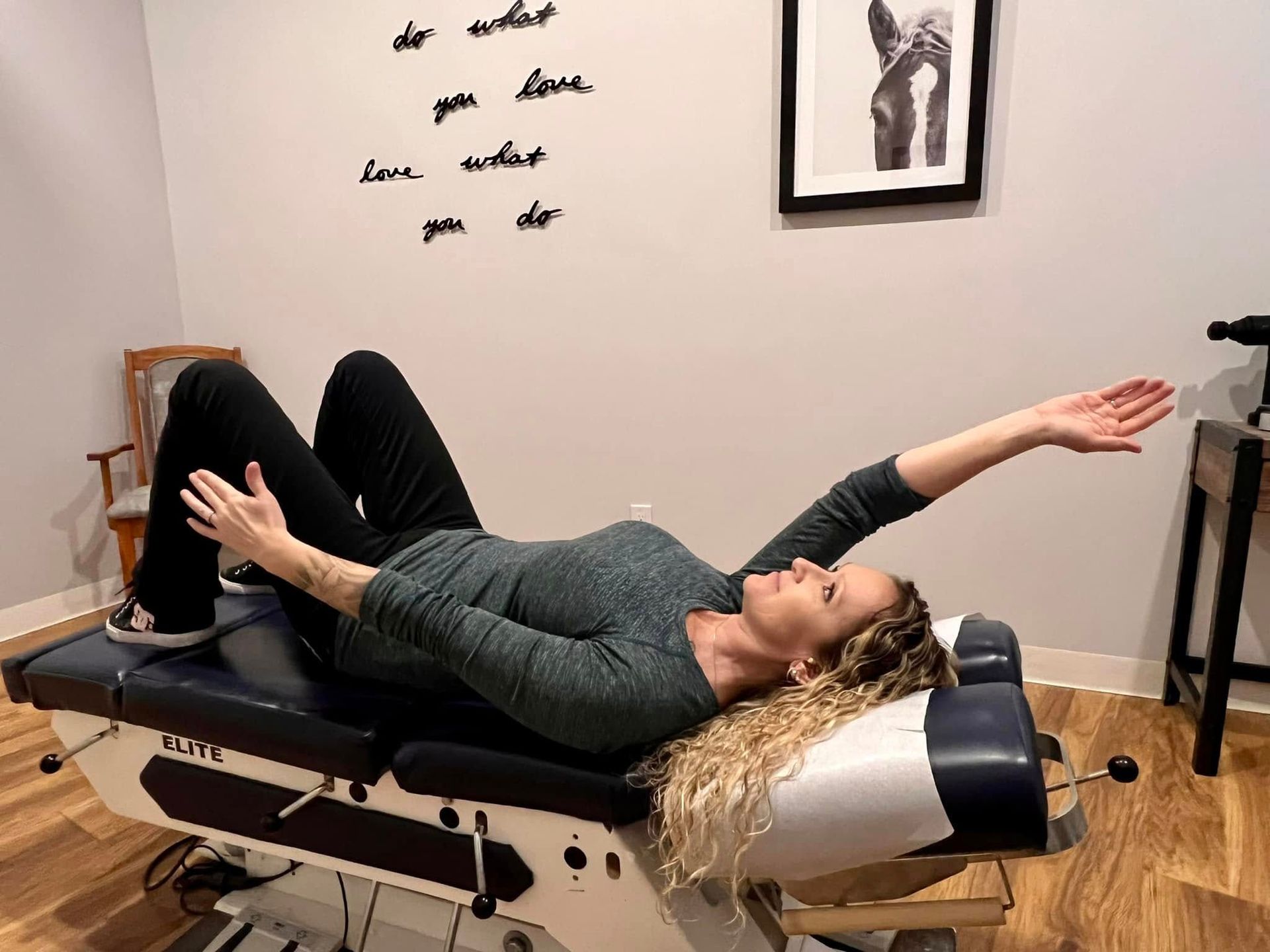A person lies on a chiropractic table, knees bent and one arm extended upward, in a clinical room.