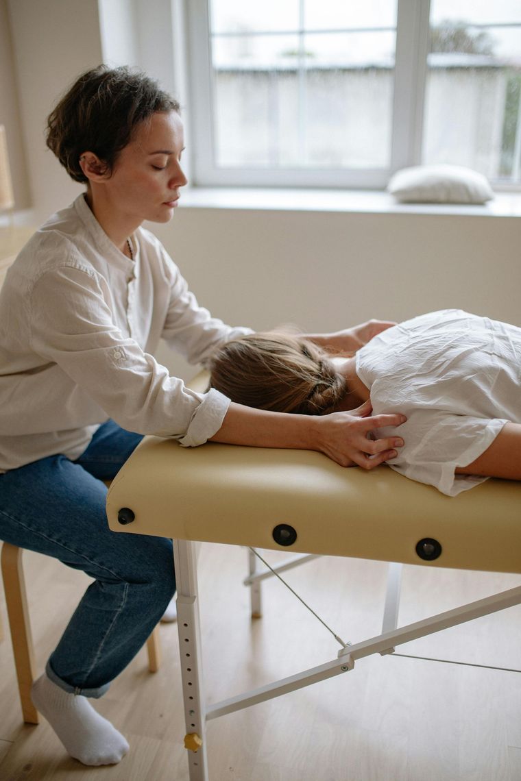 A person in a white shirt and jeans performs a massage on a patient lying on a treatment table in a bright, indoor room.
