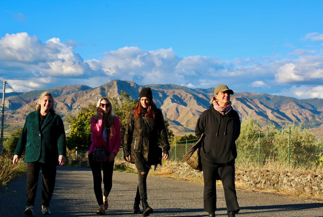 Three people walk along a stone path beside a building, with mountains in the distance.