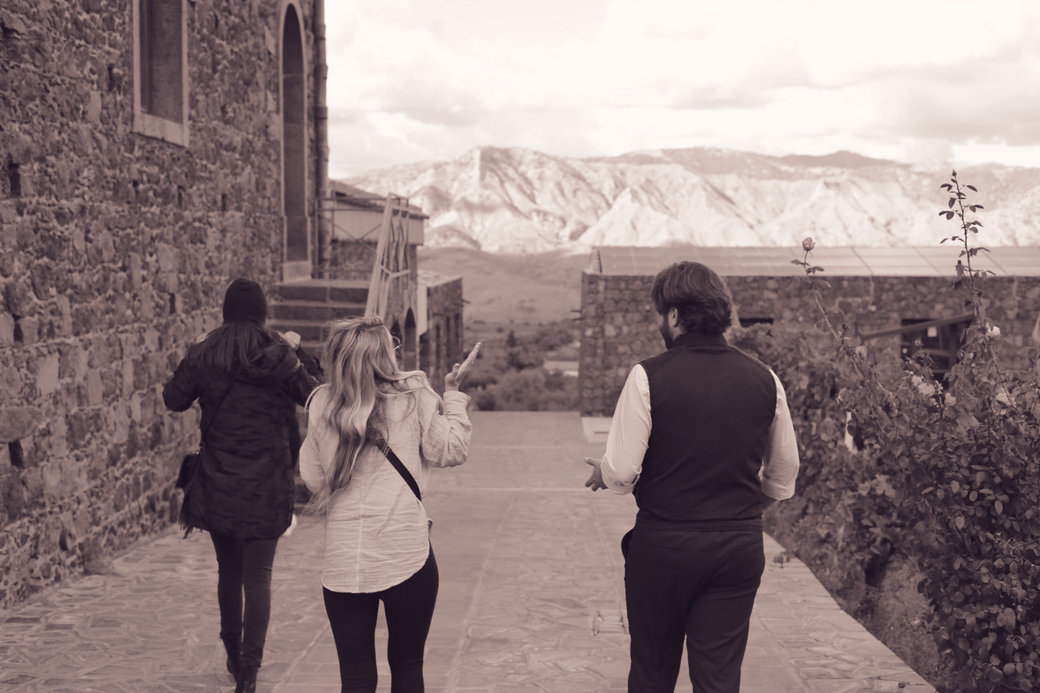 Three people walk along a stone path beside a building, with mountains in the distance.