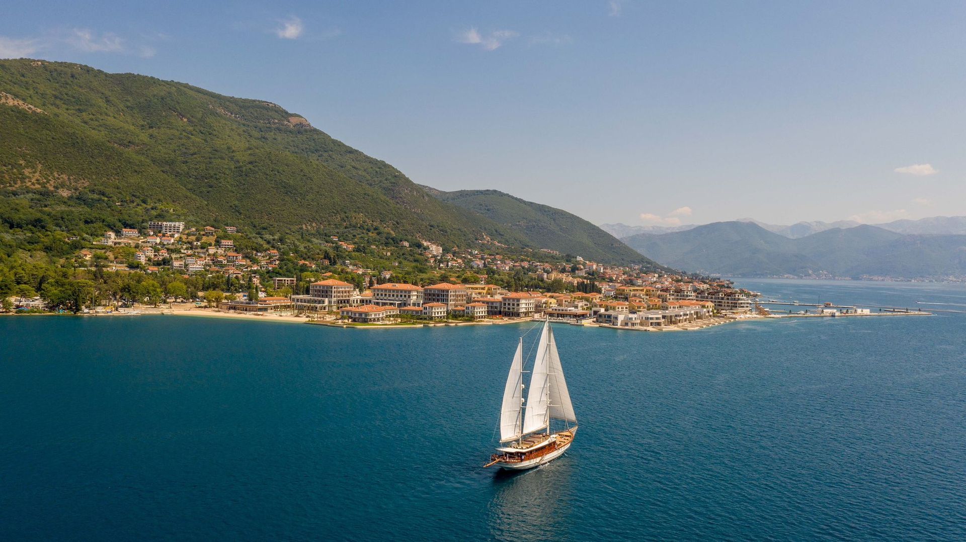Sailboat gliding on blue sea beside a coastal town and green hills under a clear sky
